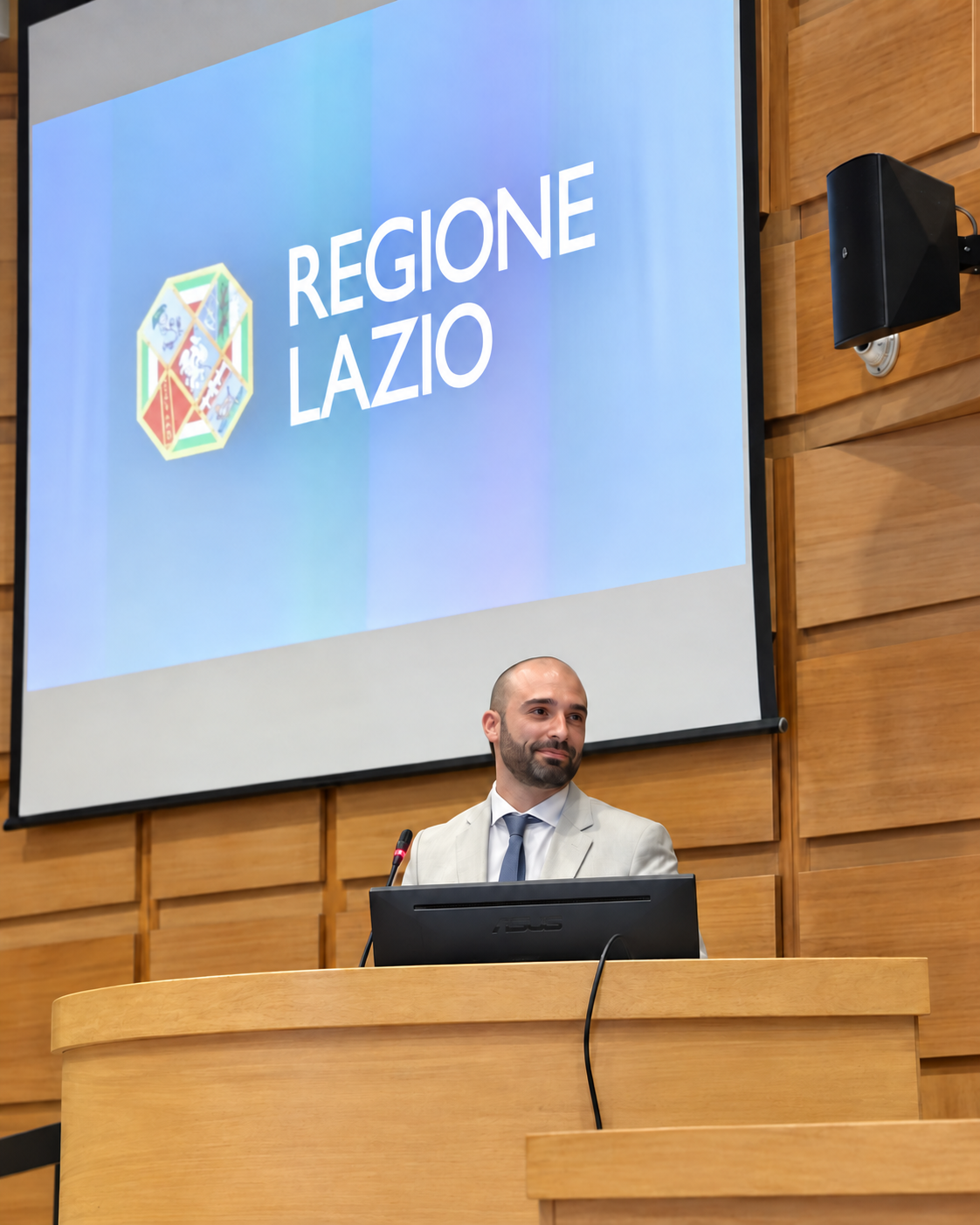 Un uomo in abito chiaro dietro un banco con microfono, davanti a uno schermo con la scritta Regione Lazio.